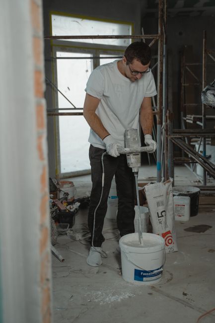 A male construction worker operating a concrete mixer during an after builders cleaning process in a renovation project at a residential property in Holland Park W8. The room is in the midst of development, with unfinished walls, visible scaffolding, and construction materials scattered on the floor. Bright natural light streams through a large window, illuminating the space. The worker, dressed in casual clothing and white gloves, focuses on pouring or mixing materials, with a white bucket and packaging of construction supplies nearby. Cleaners W8 provides comprehensive cleaning services to restore hygiene and surface cleanliness after builders work, ensuring floors, surfaces, and fixtures like wood and tiled areas are free from dust, debris, and residual materials, suitable for both domestic and commercial settings.