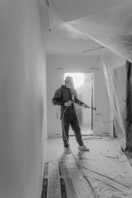A worker wearing protective gear, including a mask, gloves, and overalls, is performing surface cleaning in a room during after builders cleaning for Holland Park W8 renovations. The worker is holding a spray nozzle connected to a hose, applying a cleaning or sanitising solution to the walls and ceiling, which are temporarily covered with plastic sheeting to protect surfaces. The room features a wooden or laminate floor partially covered with a protective plastic sheet, and the space appears well-lit with natural light coming from a doorway at the far end. The overall scene showcases the thorough post-construction cleaning process carried out by Cleaners W8, focusing on deep cleaning surfaces to remove dust, construction debris, and residues to restore hygiene and shine in the residential or commercial space.