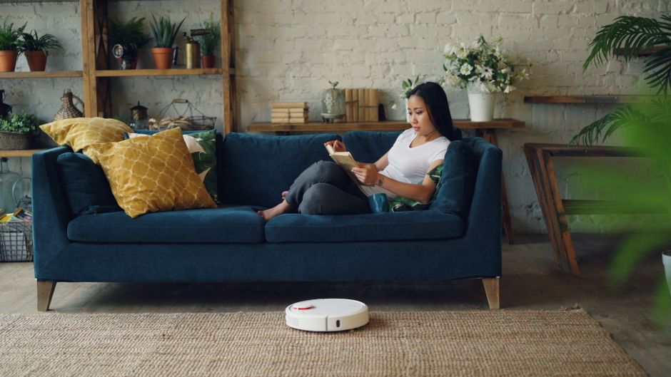 A woman sitting on a dark blue fabric sofa with wooden legs in a living room, using a tablet. The sofa is decorated with a mustard yellow textured cushion and a white cushion with a colorful pattern. The room features a light-colored exposed brick wall, wooden wall-mounted shelves with potted plants, books, and decorative objects. To the right, there is a large green leafy plant on the floor. The floor is covered with a natural woven rug. In front of the sofa, a white robotic vacuum cleaner is positioned on the rug, suggesting recent surface cleaning. The lighting is natural, coming from an unseen window, illuminating the space with a clean and tidy appearance. This scene reflects domestic surface cleaning and maintenance performed by professionals like Cleaners W8, showcasing a well-maintained, hygienic living environment.