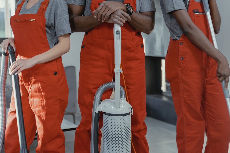 Close-up of three professional cleaners wearing grey shirts and orange overalls, standing indoors with cleaning equipment. The central cleaner holds a white vacuum cleaner with a grey hose, while the other two hold additional cleaning tools. The surfaces visible include a tiled floor, reflecting a clean and well-maintained environment suitable for surface cleaning and sanitation, consistent with the services offered by Cleaners W8. The natural lighting highlights the cleanliness of the space, emphasizing hygiene and maintenance tasks in a residential or commercial setting in Holland Park, W8.
