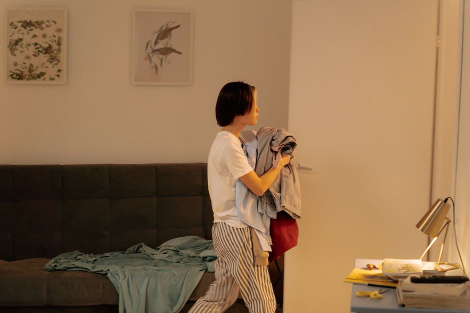 A woman sitting on a dark blue fabric sofa with wooden legs in a living room, using a tablet. The sofa is decorated with a mustard yellow textured cushion and a white cushion with a colorful pattern. The room features a light-colored exposed brick wall, wooden wall-mounted shelves with potted plants, books, and decorative objects. To the right, there is a large green leafy plant on the floor. The floor is covered with a natural woven rug. In front of the sofa, a white robotic vacuum cleaner is positioned on the rug, suggesting recent surface cleaning. The lighting is natural, coming from an unseen window, illuminating the space with a clean and tidy appearance. This scene reflects domestic surface cleaning and maintenance performed by professionals like Cleaners W8, showcasing a well-maintained, hygienic living environment.