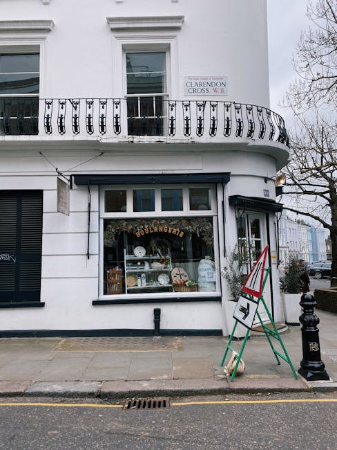 Photograph of a corner storefront with a large display window showcasing baked goods and pottery items, framed by a black metal awning. The building has a white exterior with a decorative wrought iron balcony above, and a sign indicating 'BOULANGERIE' inside the window. On the sidewalk outside, there is a green and red A-frame sign, a black bollard, and a black drainage grate visible on the pavement. The scene is captured during the day with overcast sky, and the street appears clean and well-maintained. This setting exemplifies commercial cleaning in a retail environment, highlighting the importance of surface hygiene and tidy presentation for shops in Holland Park, W8. 

Cleaners W8 specializes in comprehensive cleaning services for shops like this, ensuring surfaces such as glass, ceramic tiles, and tiled floors remain spotless and hygienic, contributing to an inviting atmosphere for customers.