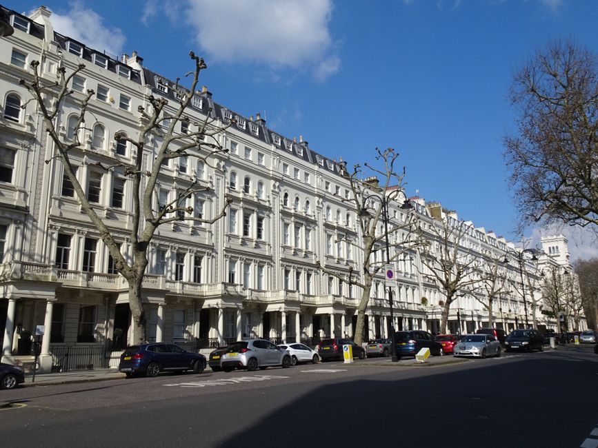 Photograph of a residential street in Holland Park, W8, showcasing elegant Victorian-style terraced flats with white facades, decorative moldings, arched windows, and black wrought iron balconies. In