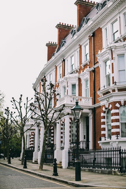 The image depicts the façade of a Victorian-style residential building with white decorative trim and red brick walls, typical of Holland Park Avenue flats in W8. The building features large sash windows with white frames, ornate moldings, and a black wrought iron fence along the sidewalk. In front, there are leafless trees planted in a row, with black street lamps positioned between them, illuminating the area during the evening. The scene is captured in natural daylight, highlighting the building's historical architectural details and well-maintained exterior. Cleaners W8 specialise in surface cleaning and deep cleaning services for such properties, ensuring high standards of hygiene and shine. Their expertise includes thorough sanitisation of external surfaces and windows, maintaining the aesthetic appeal of residential flats in Holland Park Avenue.