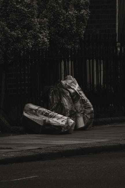 Three large black garbage bags filled with waste are placed on the sidewalk in front of a black metal fence and dense greenery. The bags appear to be made of thick plastic and are crumpled, indicating they are filled and tied. The scene is set outdoors in a residential area, with a pavement curb visible in the foreground. The lighting suggests a cloudy or overcast day. This image relates to waste management and cleanliness, which are key aspects of residential and commercial cleaning services provided by Cleaners W8, especially in accordance with Kensington and Chelsea council rules for waste in W8, W8. Proper waste disposal and maintaining clean surroundings are essential parts of surface cleaning and hygiene practices offered by professional cleaning companies.