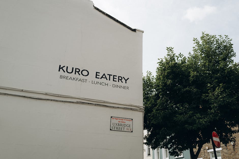 Exterior view of a white building with a sign reading 'KURO EATERY' offering breakfast, lunch, and dinner, situated on Uxbridge Street in W8. The building features a minimalistic facade with clean, smooth surfaces and has visible electrical wiring running along the wall. To the right, there is a well-maintained green tree providing natural shade, and a traffic sign indicating no entry can be seen in the background. Natural daylight illuminates the scene, highlighting the neatness and clarity of the signage and the surrounding environment. This image is associated with Cleaners W8, emphasizing the importance of maintaining clean, hygienic surfaces in commercial spaces such as eateries, especially in accordance with local waste and sanitation regulations.