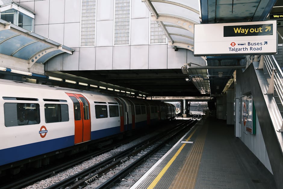 A subway train with a white, orange, and blue exterior is stopped at a modern underground station platform near Holland Park tube station, W8. The platform surface is clean and features yellow tactile paving strips along the edge for safety. Overhead, a curved glass canopy provides shelter, allowing natural light to partially illuminate the area. Signage indicates directions for exits and bus connections, with a prominent yellow 'Way out' sign and additional signs for bus routes towards Talgarth Road. The station's walls are made of grey and silver panels, contributing to a sleek, urban appearance. Cleaners W8 specializes in surface cleaning, sanitisation, and deep cleaning services, ensuring hygienic and well-maintained environments in residential and commercial spaces near Holland Park, and their work is reflected in the tidy and pristine station surroundings.