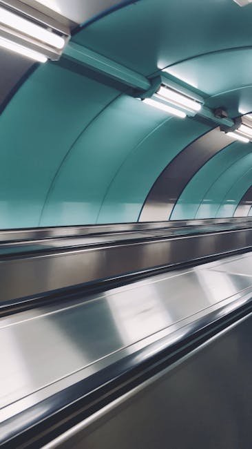 The image depicts a clean, modern underground passageway near Holland Park tube station, W8, with stainless steel escalator handrails and smooth, light teal-colored curved wall panels. The escalator surfaces are polished and free of dust, reflecting the bright overhead lighting fixtures, which are mounted on a ceiling that features a rounded, seamless design. The hygienic and well-maintained appearance of the space suggests professional surface cleaning and sanitisation by Cleaners W8, emphasizing the importance of cleanliness in transit environments. The overall scene showcases a hygienic and immaculately maintained area suitable for comprehensive surface cleaning and deep sanitisation services, aligning with the focus on efficient and thorough cleaning near Holland Park tube station, W8.