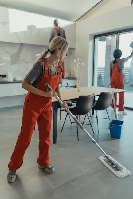 A female cleaner from Cleaners W8 wearing red overalls and a grey shirt is performing surface cleaning in a modern kitchen area. She is using a flat mop on the large, smooth, grey tiled floor, which appears clean and dry. In the background, two other cleaners, also in red overalls, are working near a marble kitchen counter with a white and grey finish, and wooden accents. The kitchen features a large window allowing natural light to illuminate the space, highlighting the shiny, polished surfaces and the glass dining table surrounded by black chairs. A blue cleaning bucket is positioned on the floor nearby. The scene emphasizes thorough domestic cleaning and surface maintenance in a contemporary residential setting, with a focus on hygiene and cleanliness, reflecting the services offered by Cleaners W8 for top-rated cleaning in Holland Park W8.