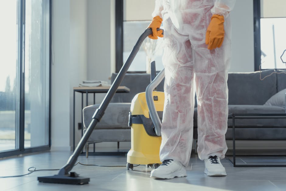 A female cleaner from Cleaners W8 wearing red overalls and a grey shirt is performing surface cleaning in a modern kitchen area. She is using a flat mop on the large, smooth, grey tiled floor, which appears clean and dry. In the background, two other cleaners, also in red overalls, are working near a marble kitchen counter with a white and grey finish, and wooden accents. The kitchen features a large window allowing natural light to illuminate the space, highlighting the shiny, polished surfaces and the glass dining table surrounded by black chairs. A blue cleaning bucket is positioned on the floor nearby. The scene emphasizes thorough domestic cleaning and surface maintenance in a contemporary residential setting, with a focus on hygiene and cleanliness, reflecting the services offered by Cleaners W8 for top-rated cleaning in Holland Park W8.